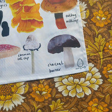 Load image into Gallery viewer, A close up of the bottom right of the off white tea towel, showing the duck egg designs logo in black on the bottom right. Next to the logo you can see a charcoal burner and an ink cap, above which is a chicken of the wood and an oak bug milkcap. To the right and below the teatowel you can see a warm brown retro fabric with a floral pattern.
