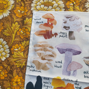 A close up of the top left of the tea towel showing some of the illustrations in more detail. From L to R, top to bottom you can see a velvet shank, a shaggy parasol, and oyster mushroom and a wood blewit fungi, all on an off white background. Behind the tea towel to the top and left you can see a warm brown floral patterned retro fabric in the background.