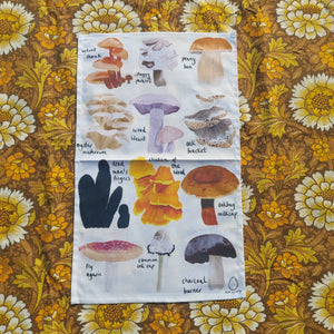 An off white tea towel featuring 12 different British fungi in rows of three with their names in black handwriting next to them. Behind the tea towel is a warm brown retro floral patterned fabric.