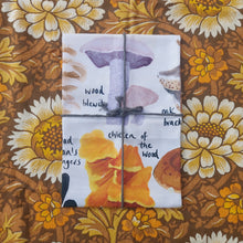 Load image into Gallery viewer, A white tea towel featuring a range of different fungi sits folded and tied in grey macrame twine. Behind the tea towel you can see a warm brown retro floral patterned fabric. 
