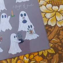 Load image into Gallery viewer, A close up of the dusky purple print showing the group of ghosts in the bottom right. Below and to he right side of the print you can see a warm brown retro floral patterned fabric.
