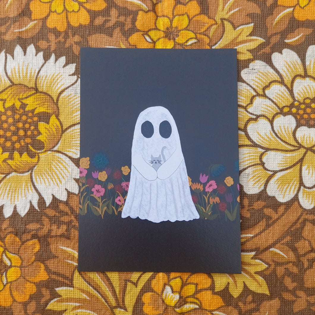 A black print featuring a white ghost holding a little grey cat while standing in a field of autumnal flowers. Behind the print you can see a warm brown retro floral patterned fabric.