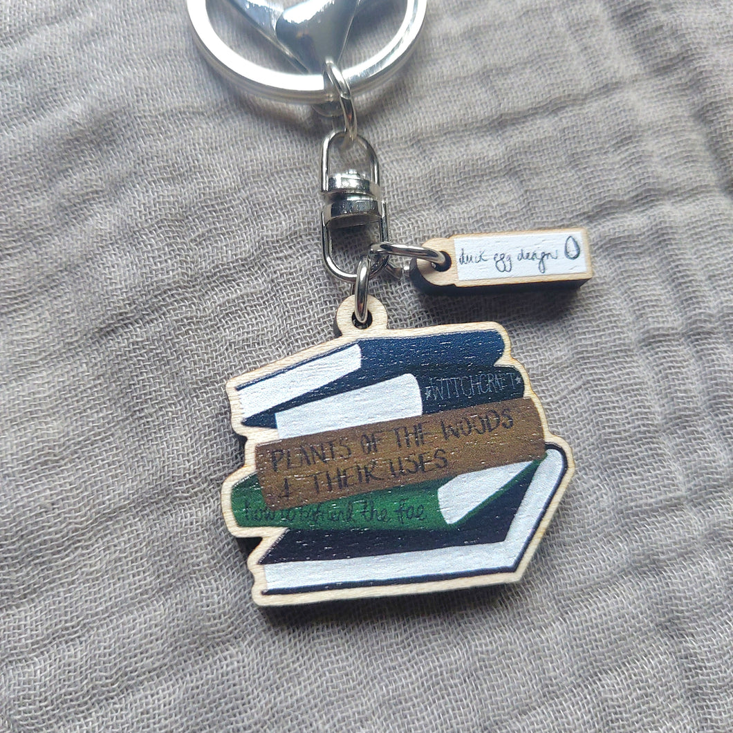 A wooden key ring lies on an off grey fabric background. The key ring features a wooden charm with a stack of books in different colours.
