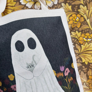 A close up view of the ghost holding the grey kitten on a black background with some pink, warm yellow, red and blue flowers visible at the bottom. The print is on a white tote bag which lies on a warm brown floral patterned background.