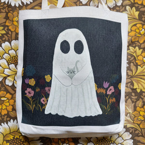 A close up of view of a white tote with a black square printed on it featuring a white ghost holding a grey kitten amid a row of pink, red and mustard yellow colours. Behind the tote you can see a warm brown floral retro patterned fabric.