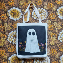 Load image into Gallery viewer, A white textured tote bag sits in the middle of the image on a brown retro floral patterned fabric. The tote has a black square printed on it featuring a white ghost holding a grey kitten amid a row of pink, red and mustard yellow colours.
