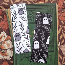 Load image into Gallery viewer, An image of the front and back of the bookmark together showing the black and white versions of the ghost and vine pattern. Behind the bookmarks you can see an old green gardening book behind which is a brown floral patterned fabric.
