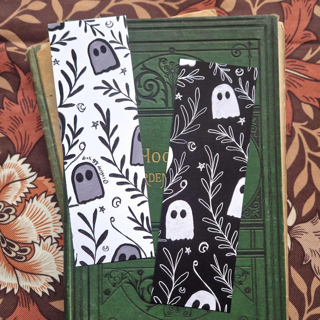 An image of the front and back of the bookmark together showing the black and white versions of the ghost and vine pattern. Behind the bookmarks you can see an old green gardening book behind which is a brown floral patterned fabric.