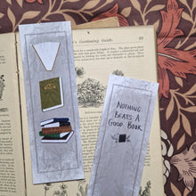 Load image into Gallery viewer, Two bookmarks sit in an open vintage gardening book showing the front and back of the bookmark. Behind the book you can see a brown retro floral fabric. The bookmark has a textured grey background with a dark grey border and leafy vines pattern. Running down the front of the bookmark are three different book illustrations while on the back there are words in black handwriting that say ‘Nothing beats a good book’ at the bottom of the bookmark is the duck egg designs logo.

