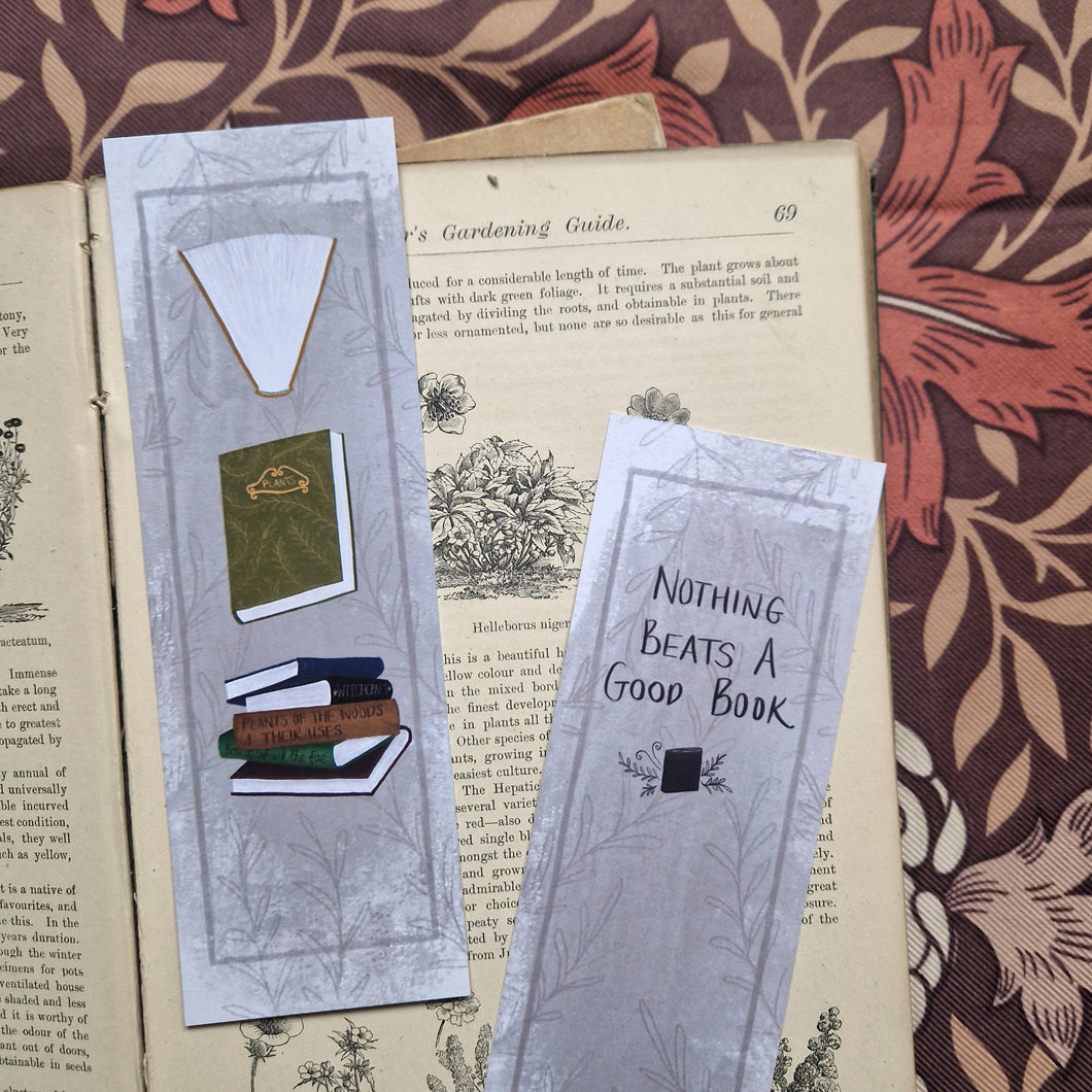 Two bookmarks sit in an open vintage gardening book showing the front and back of the bookmark. Behind the book you can see a brown retro floral fabric. The bookmark has a textured grey background with a dark grey border and leafy vines pattern. Running down the front of the bookmark are three different book illustrations while on the back there are words in black handwriting that say ‘Nothing beats a good book’ at the bottom of the bookmark is the duck egg designs logo.
