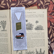 Load image into Gallery viewer, The front of the bookmark is visible in an open vintage gardening book on a brown retro floral patterned fabric. The bookmark is a textured grey with a darker grey border line running around the outside as well as a leafy vine design. Running down the bookmark are three different book illustrations - a purple book, a stack of books and then a green book. 
