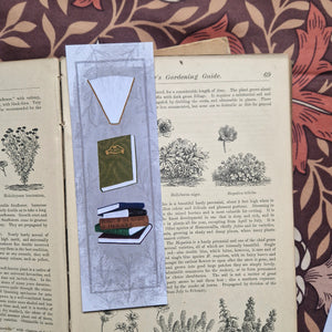 The front of the bookmark is visible in an open vintage gardening book on a brown retro floral patterned fabric. The bookmark is a textured grey with a darker grey border line running around the outside as well as a leafy vine design. Running down the bookmark are three different book illustrations - a purple book, a stack of books and then a green book. 