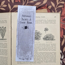 Load image into Gallery viewer, The back of the bookmark is on display showing the words ‘Nothing beats a good book’ in black handwriting on the textured grey background. There are simple leafy vines in the background and a deep grey border line. Behind the bookmark is an open vintage gardening book on a brown retro floral background.
