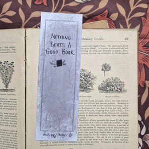 The back of the bookmark is on display showing the words ‘Nothing beats a good book’ in black handwriting on the textured grey background. There are simple leafy vines in the background and a deep grey border line. Behind the bookmark is an open vintage gardening book on a brown retro floral background.