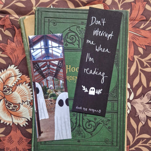 This image shows the front and back of the bookmark next to each other sat on a vintage green book. The left bookmark shows a portion of a greenhouse with several ghosts in it; the right bookmark is black with white writing on it that reads ‘Don’t interrupt me when I’m reading’ in white above a white ghost with leafy vines around it with a duck egg designs logo at the bottom. Surrounding the book you can see a brown floral background.