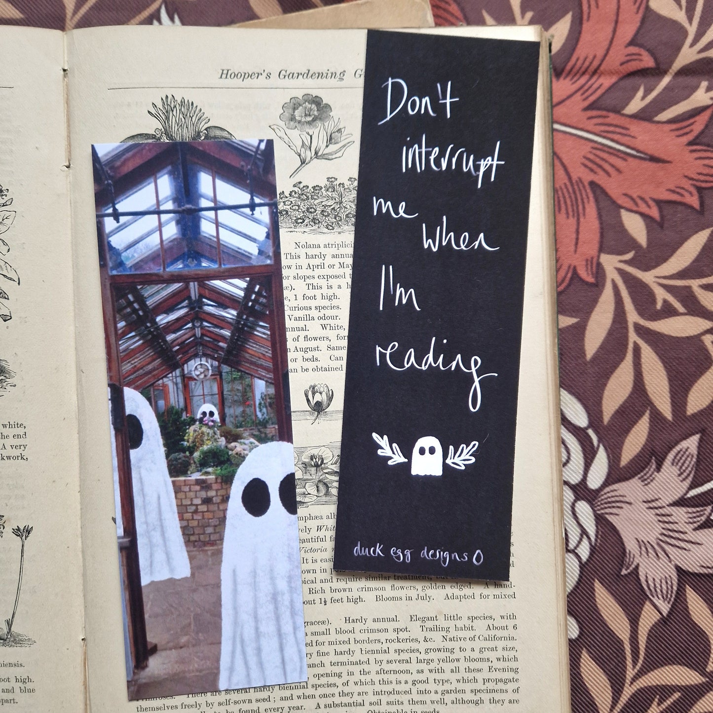 An old book sits open with a brown floral background visible to the right and top of it. The book has two bookmarks on it - one features a greenhouse with ghosts in it while the other is black and features the words ‘Don’t interrupt me when I’m reading’ in white above a ghost with little leafy vines around it. At the bottom of the bookmark you can see a white duck egg designs logo.