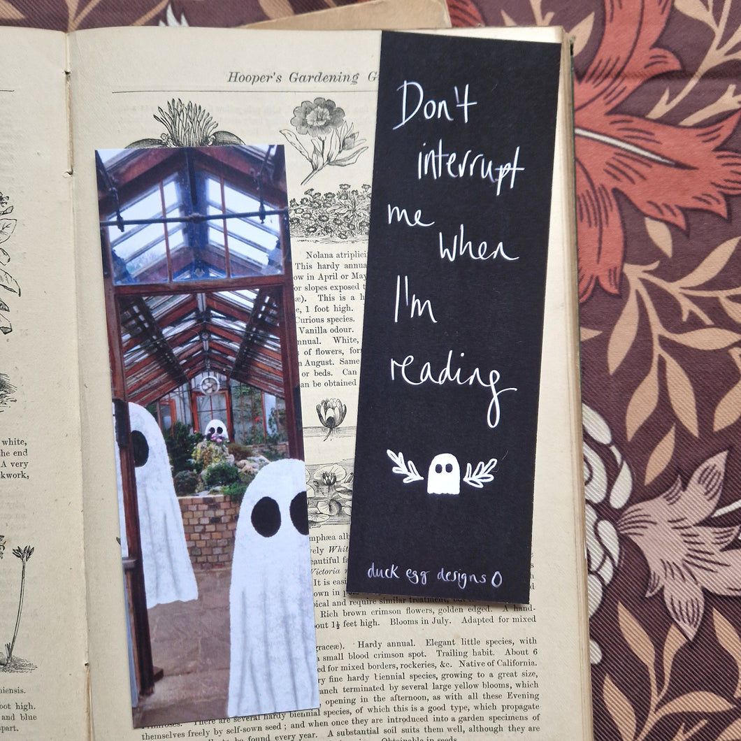 An old book sits open with a brown floral background visible to the right and top of it. The book has two bookmarks on it - one features a greenhouse with ghosts in it while the other is black and features the words ‘Don’t interrupt me when I’m reading’ in white above a ghost with little leafy vines around it. At the bottom of the bookmark you can see a white duck egg designs logo.