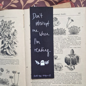 An old book lies open with a black bookmark in it with words that say ‘Don’t interrupt me when I’m reading’ in white handwriting above -a ghost with leafy vines around it. At the bottom of the bookmark is a duck egg designs logo, and behind the book you can see a brown floral background.