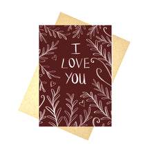 Load image into Gallery viewer, A deep red card sits on a brown envelope in front of a white background. The card features the words ‘I LOVE YOU’ in white with a leafy vine and heart design growing in towards the words from the edges.
