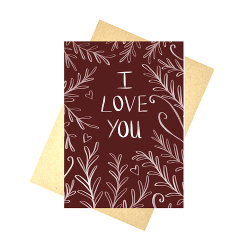 A deep red card sits on a brown envelope in front of a white background. The card features the words ‘I LOVE YOU’ in white with a leafy vine and heart design growing in towards the words from the edges.