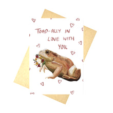 Load image into Gallery viewer, A white card sits on a brown recycled paper envelope in front of a white background. The card is covered in pink heart outlines with the words ‘Toad-ally In Love With You’ in pink handwriting above a realistic illustration of a toad with a bunch of flowers.

