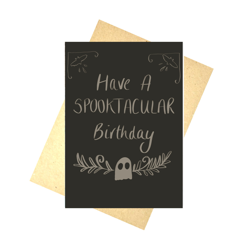A dark grey card featuring off white handwriting that reads ‘Have A SPOOKTACUALR Birthday’ above a small ghost with leafy vines growing to the sides from behind it. Above the words to the right and left of the card are bat shapes with patterns around them. Behind the card is a brown envelope behind which is a white background.