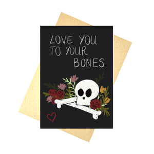 A black card sits on a brown recycled paper envelope in front of a white background. The card features the words ‘Love You To Your Bones’ in white above a skull and a couple of bones surrounded by flowers with a red outline of a heart to the bottom left of the card.