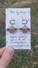 Load and play video in Gallery viewer, A video of the painted lady butterfly earrings which can be seen hanging from a white backing card with a black leafy vine design. Behind the backing card you can see a grassy background.
