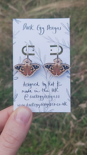 A video of the painted lady butterfly earrings which can be seen hanging from a white backing card with a black leafy vine design. Behind the backing card you can see a grassy background.