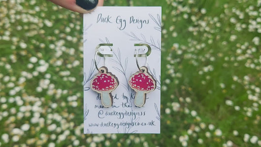 Silver hoops with red and white wooden toadstool charms on a white backing card in front of a sea of daisies.
