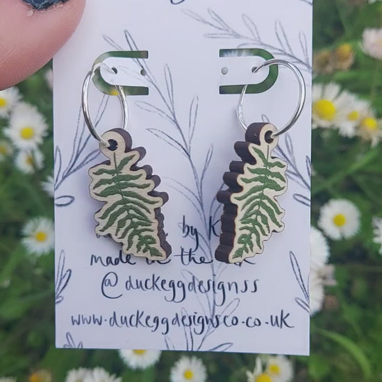 Green fern fronds on wooden charms hang from silver hoops on a white backing card in front of a sea of grass and daisies.