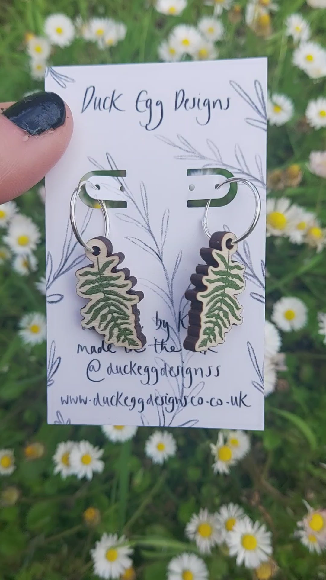 Green fern fronds on wooden charms hang from silver hoops on a white backing card in front of a sea of grass and daisies.