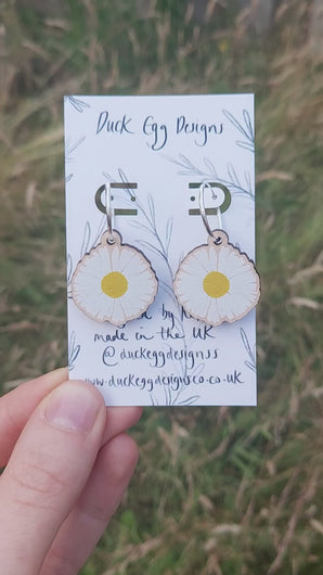 A video of silver hoops with daisy charms hanging from them on a white backing card in front of a grassy background.