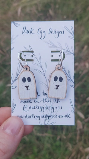 Video of silver hoops with ghost charms hanging from them. The ghosts are white with big black eyes and are hugging a dark red heart. Behind the backing card you can see a grassy background.