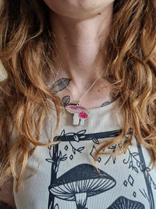 The red and white fly agaric necklace with a silver chain sits just below the collarbones of a woman with long ginger hair wearing a cream coloured top with a mushroom design.