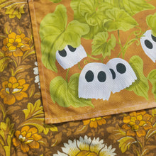 Load image into Gallery viewer, The bottom left of the tea towel is on show, with little ghost pumpkins on a green leafy vine on a warm orange background. Below and to the left of the tea towel you can see a warm brown retro floral fabric.
