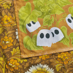 The bottom left of the tea towel is on show, with little ghost pumpkins on a green leafy vine on a warm orange background. Below and to the left of the tea towel you can see a warm brown retro floral fabric.
