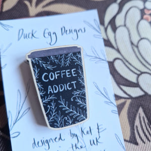 A black coffee cup wooden pin badge with the words ‘Coffee Addict’ in white handwriting and a leafy vine design sits on a white and black backing card. Behind the backing card you can see a retro brown floral patterned fabric.