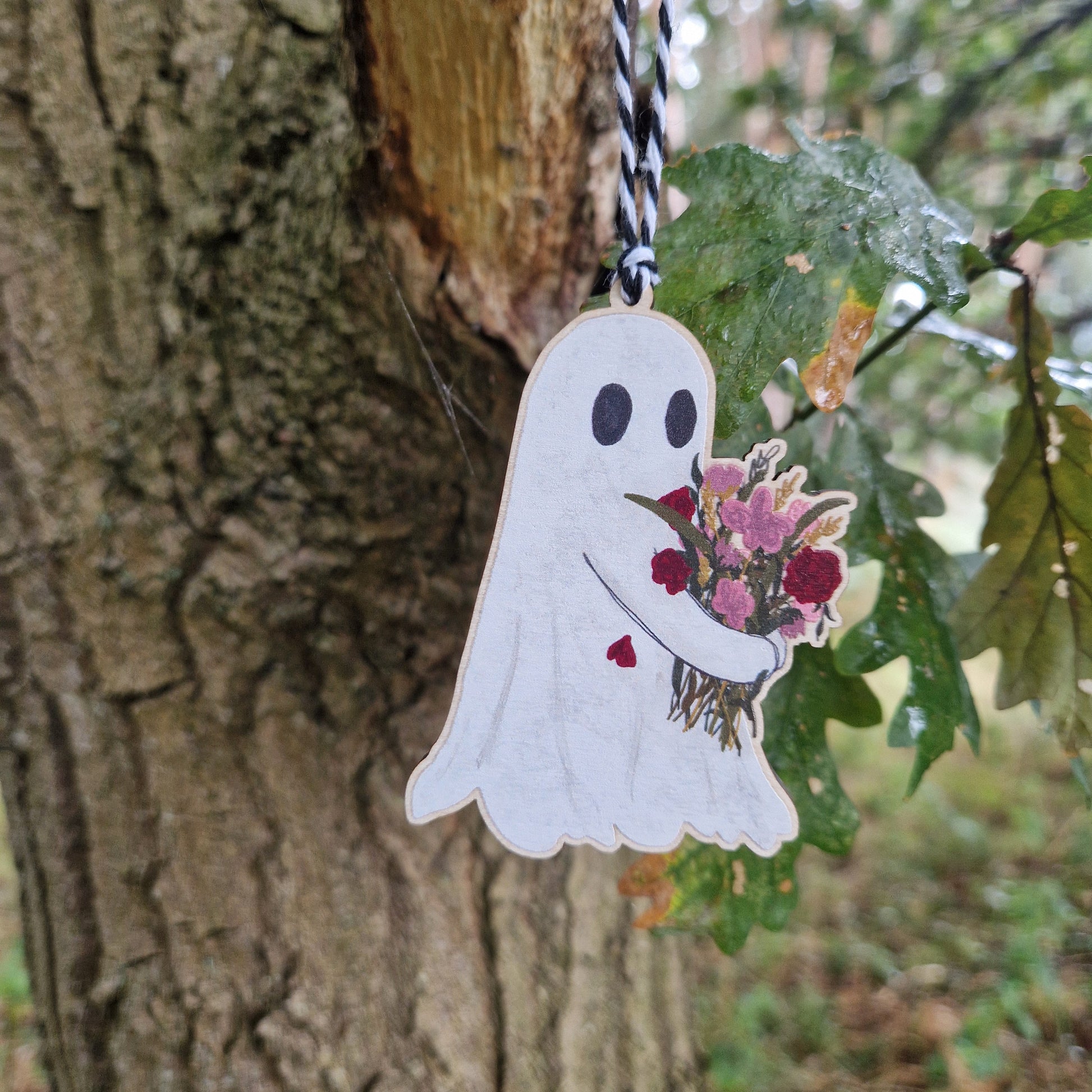 The Flower Ghost Hanging Wooden Decoration by Duck Egg Designs features a ghost holding a colorful bouquet, illustrated on wood and suspended by a string from a tree branch with green leaves.