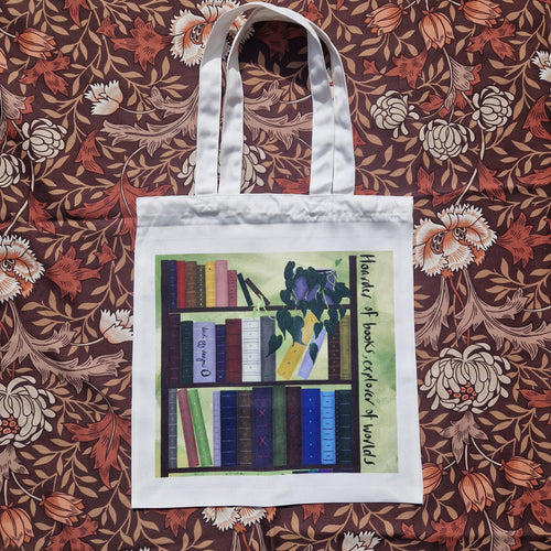 A white tote lies on a brown retro floral patterned background. The bag features a book shelf with colourful books and a trailing philodendron in front of a green textured background. To the right of the bookcase are the words ‘Hoarder of books, explorer of worlds’ in black handwriting running down the side. Across the spine of one of the books is the black Duck Egg Designs logo.