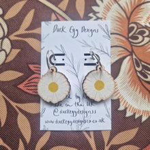 Load image into Gallery viewer, A close up of the daisy earrings with silver hoop findings demonstrating the detail in the wooden charms. The hoops sit on a white blacking card with a black leafy vine design and the words ‘Duck Egg Designs’ across the top. Behind the backing card you can see a brown and orange retro floral patterned fabric.
