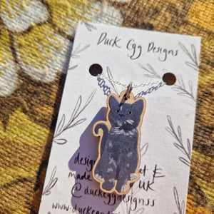 A grey cat charm sits hanging from a silver chain on a white backing card featuring a black leafy vine design. Behind the backing card you can see a warm brown retro floral fabric.