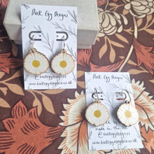 Load image into Gallery viewer, Two pairs of earrings with wooden daisy charms sit on white and black backing cards. The pair to the left is slightly higher up in the image and leans against a cream coloured box. The pair in the right has silver hoops and the backing card reads ‘Duck Egg Designs’ in black handwriting. Behind the backing cards you can see a retro floral patterned background in oranges and browns.
