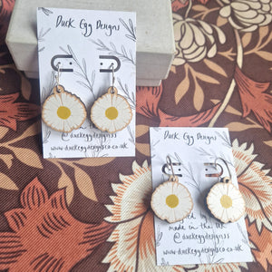 Two pairs of earrings with wooden daisy charms sit on white and black backing cards. The pair to the left is slightly higher up in the image and leans against a cream coloured box. The pair in the right has silver hoops and the backing card reads ‘Duck Egg Designs’ in black handwriting. Behind the backing cards you can see a retro floral patterned background in oranges and browns.