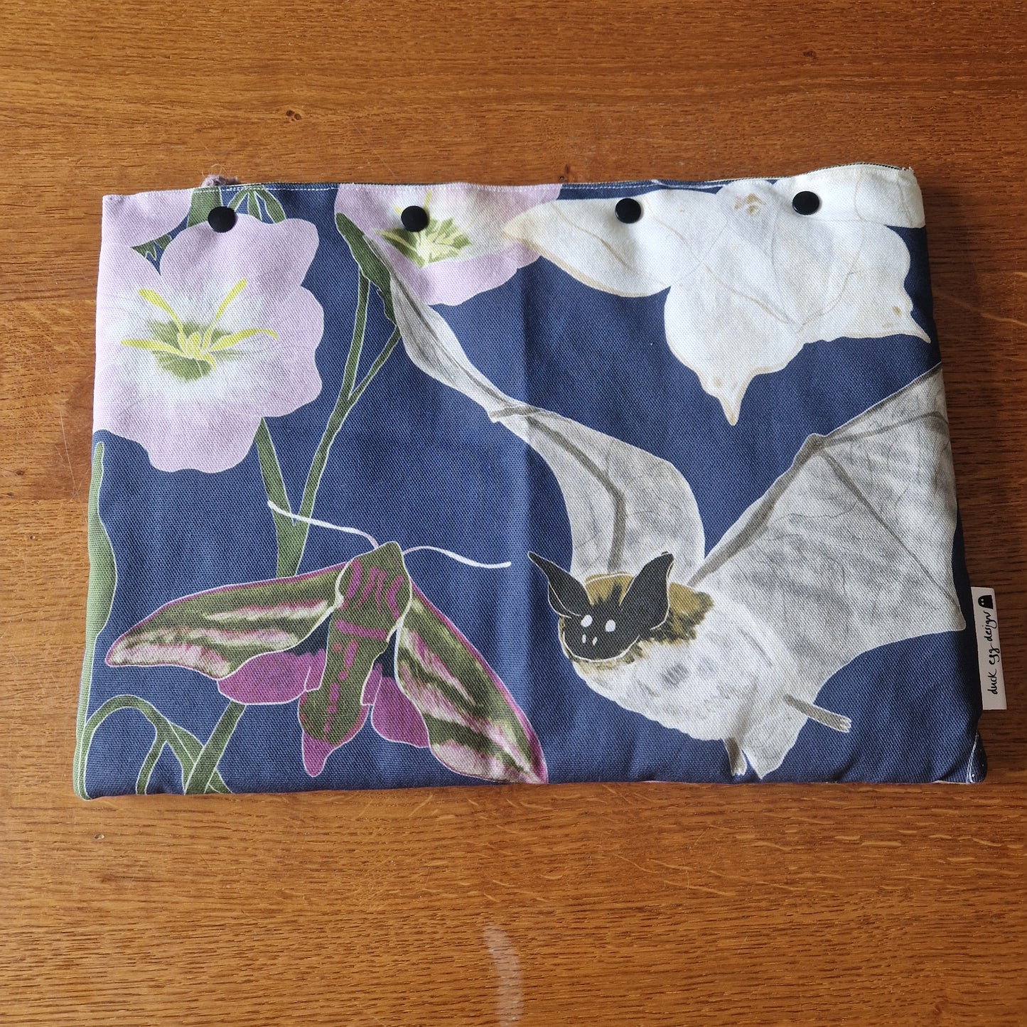 The Nocturnal Pollinators Small Laptop/Tablet Sleeve by Duck Egg Designs is a handmade pouch from recycled fabric, featuring bats, moths, and pink and white flowers on dark blue. Shown resting on a wooden surface.