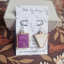 Load image into Gallery viewer, A pair of earrings featuring mismatched book charms sits on a white and black backing card which leans on a cream coloured box. The left earring has a purple book with a white leafy design and the word ‘BOTANICALS’, while the book on the right has a brown cover and sits on its spine with the pages fanning out. Behind the backing card you can see a brown retro floral background. 
