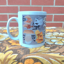 Load image into Gallery viewer, The left hand side of a white mug with a british fungi design, sat on a brown retro floral fabric in front of a red brick wall.
