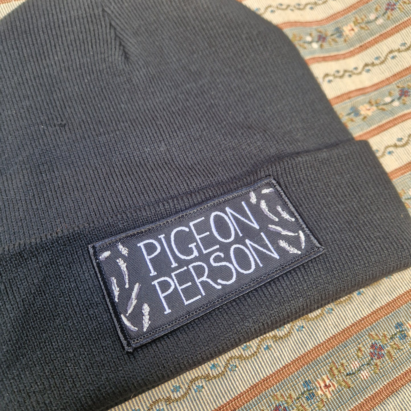 The Pigeon Person Beanie by Duck Egg Designs is a black recycled polyester hat featuring a pigeon patch with "PIGEON PERSON" in white, surrounded by feather illustrations, displayed on a patterned fabric background.