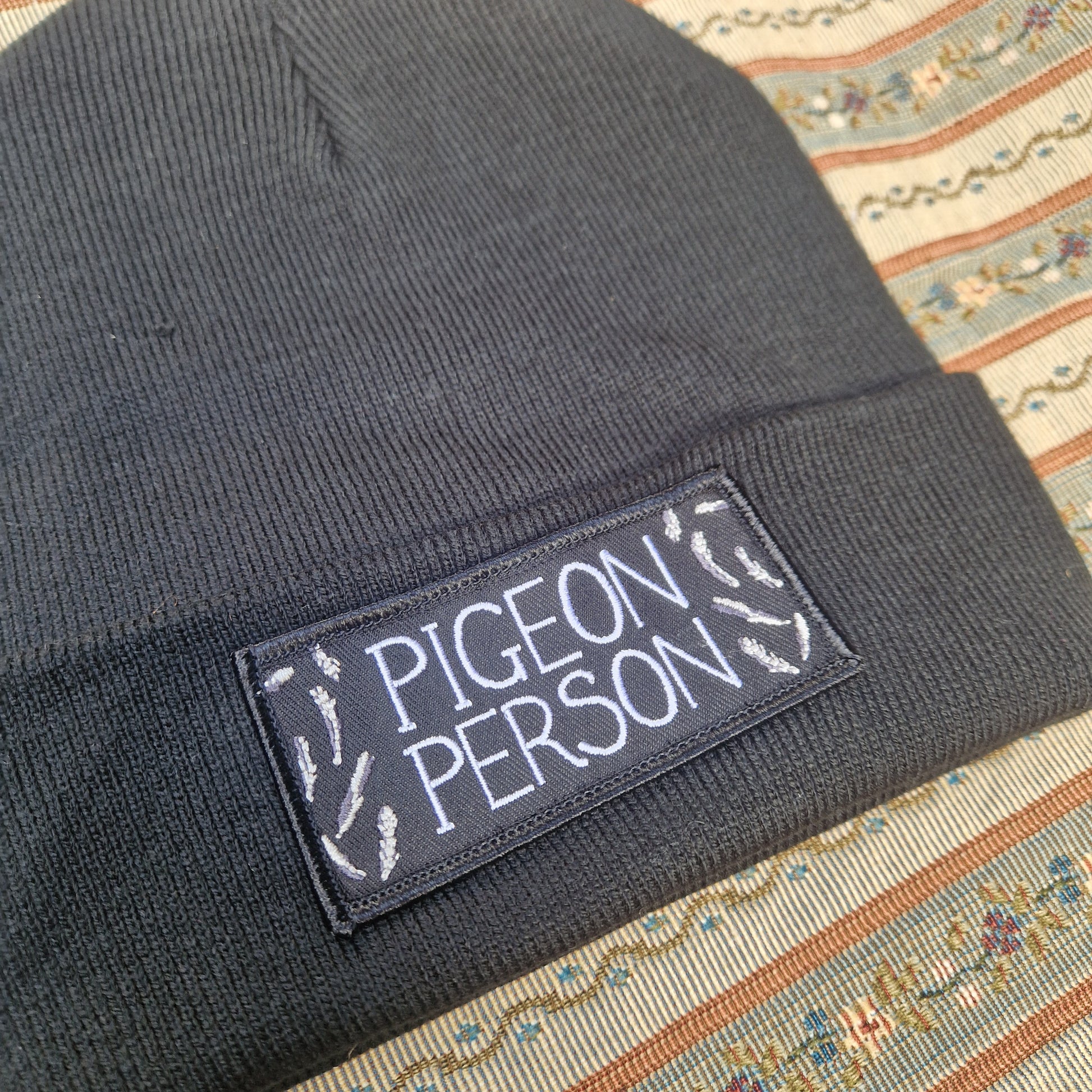 The Pigeon Person Beanie by Duck Egg Designs is a black recycled polyester hat featuring a pigeon patch with "PIGEON PERSON" in white, surrounded by feather illustrations, displayed on a patterned fabric background.