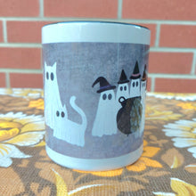 Load image into Gallery viewer, The middle section of the mug is on display showing the ghosts in that section of the mug, the mug is mainly white with a black inside and rim. The mug sits on a warm brown floral patterned fabric in front of a red brick wall.
